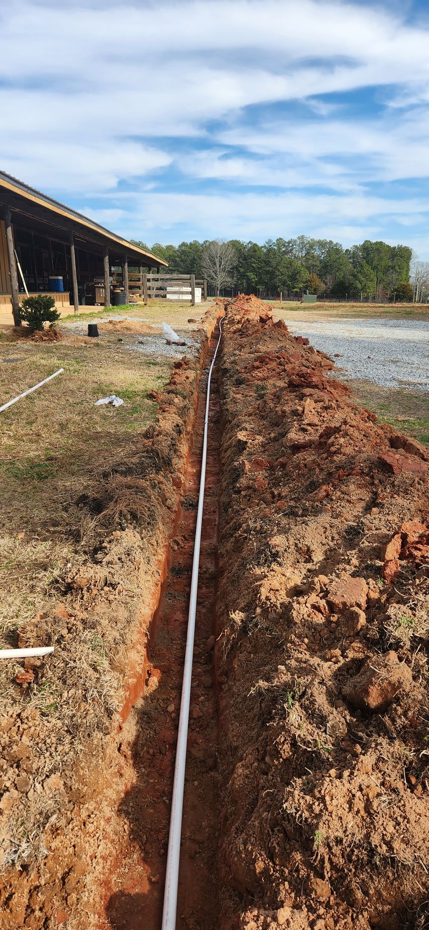 A trench with white PVC pipe is dug in red clay, near a barn under a cloudy blue sky.
