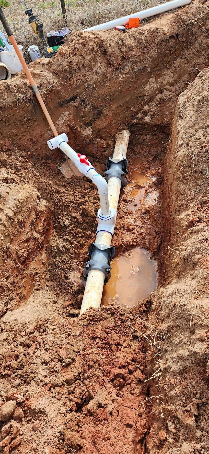Plumbing repair in a dirt trench: white pipe with fittings connected to a brown pipe.