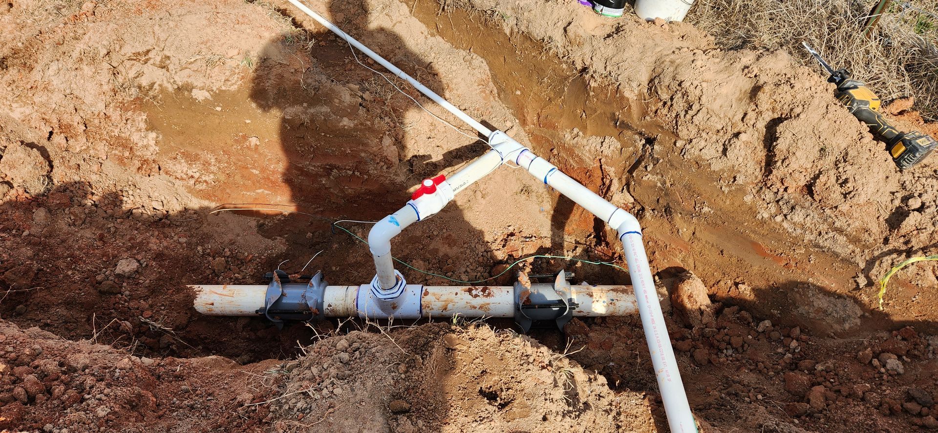 A network of white pipes with a red valve, buried in brown dirt, possibly for irrigation. A shadow of a person is visible.