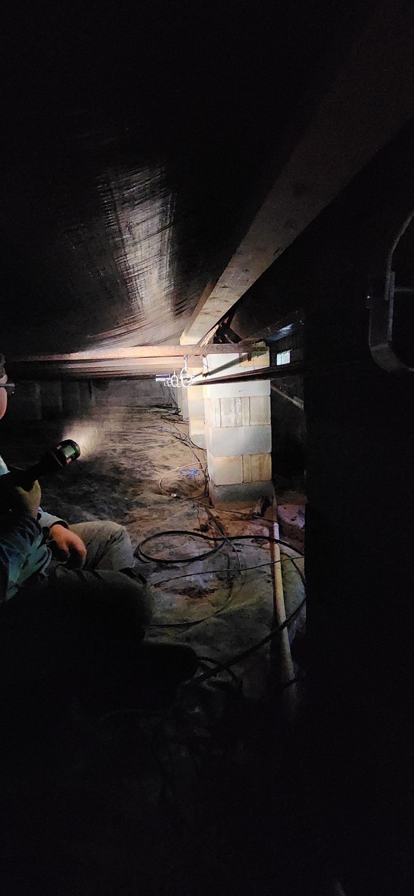 Dark, low-angle view of a crawl space. Beams, posts, and a person with a flashlight are visible.