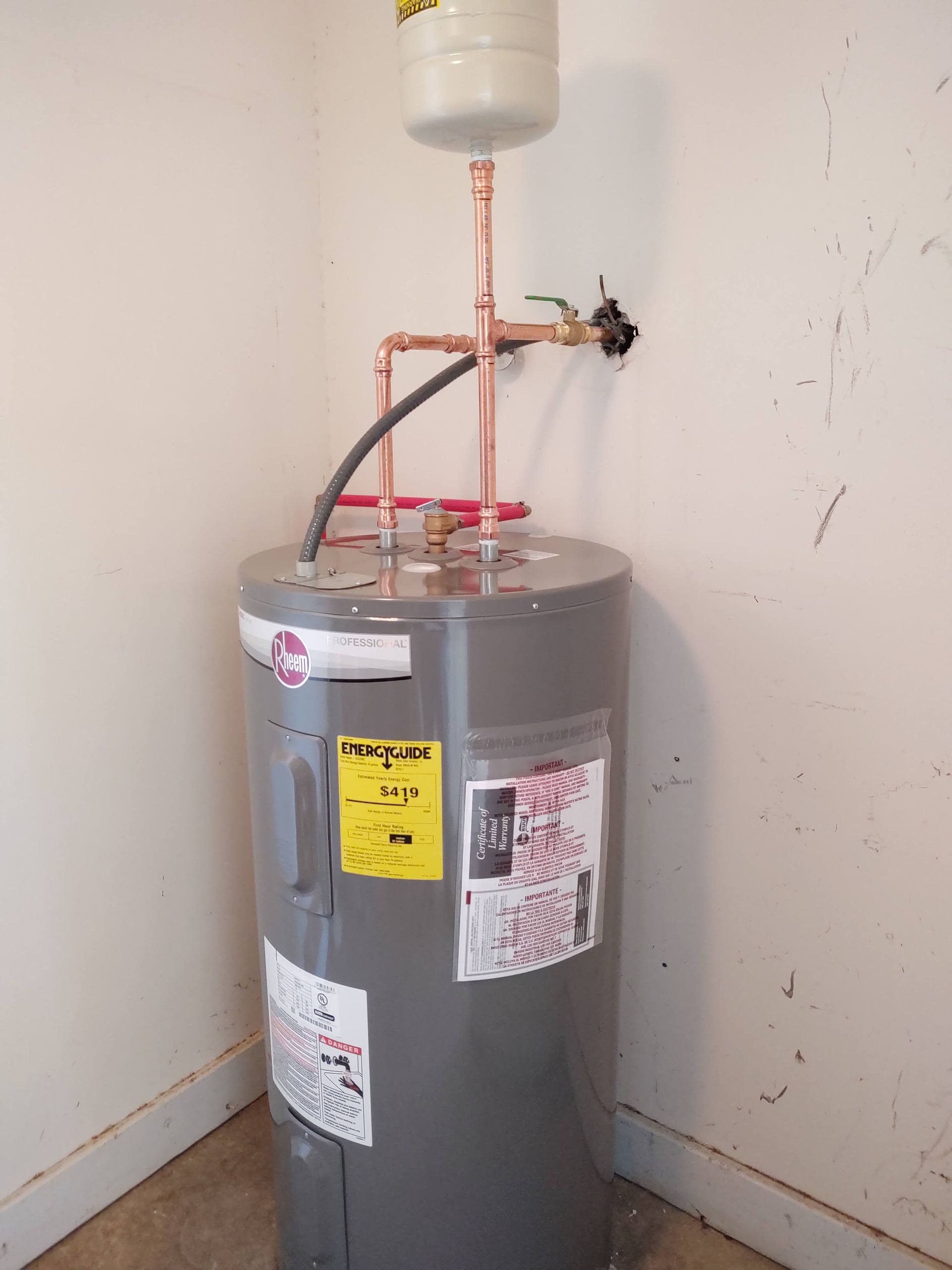 A tall gray water heater in a corner, with copper piping and a white expansion tank on top.