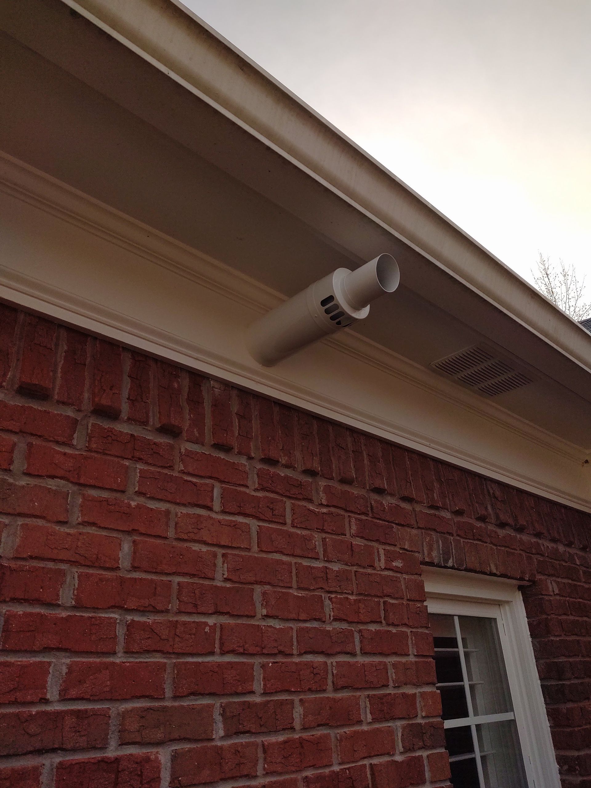 A white PVC pipe vent installed under the eaves of a red brick building.