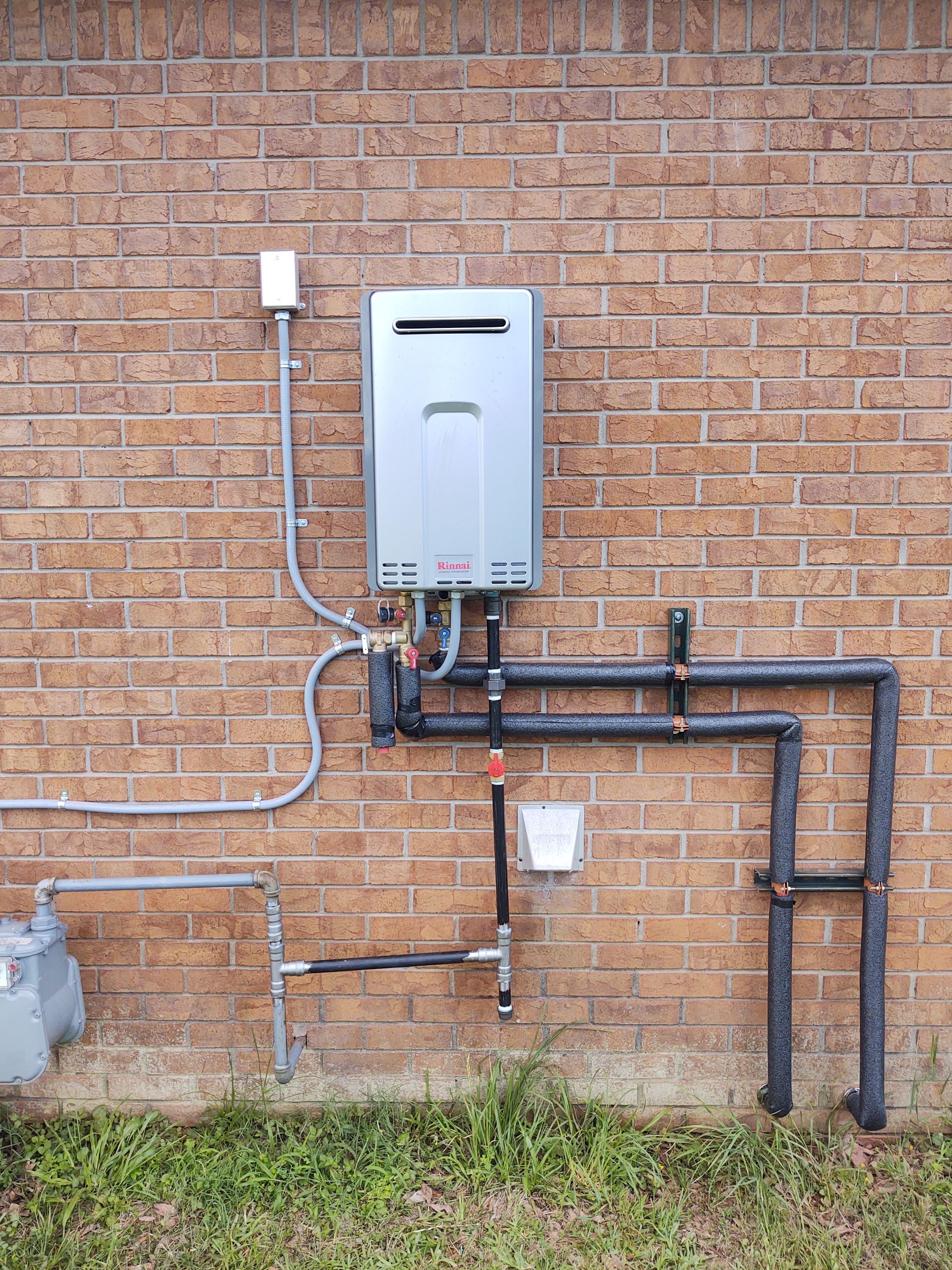 Gas-powered, tankless water heater mounted on a brick wall with connected pipes, electrical, and gas lines.