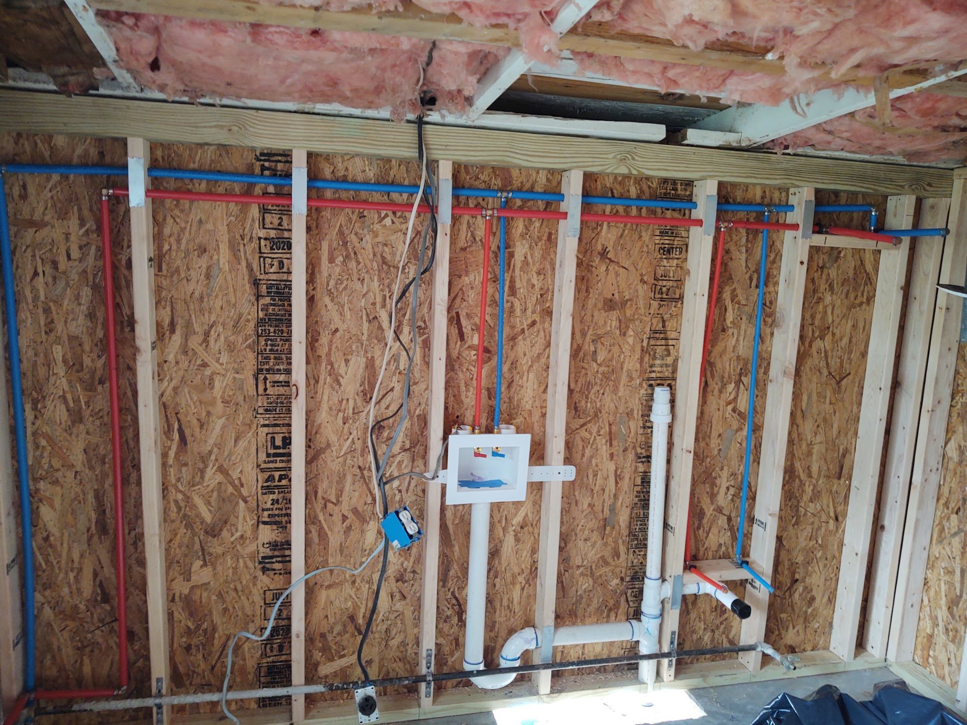 Plumbing and electrical rough-in: red/blue water pipes, electrical box, and drain pipes in a framed wall.