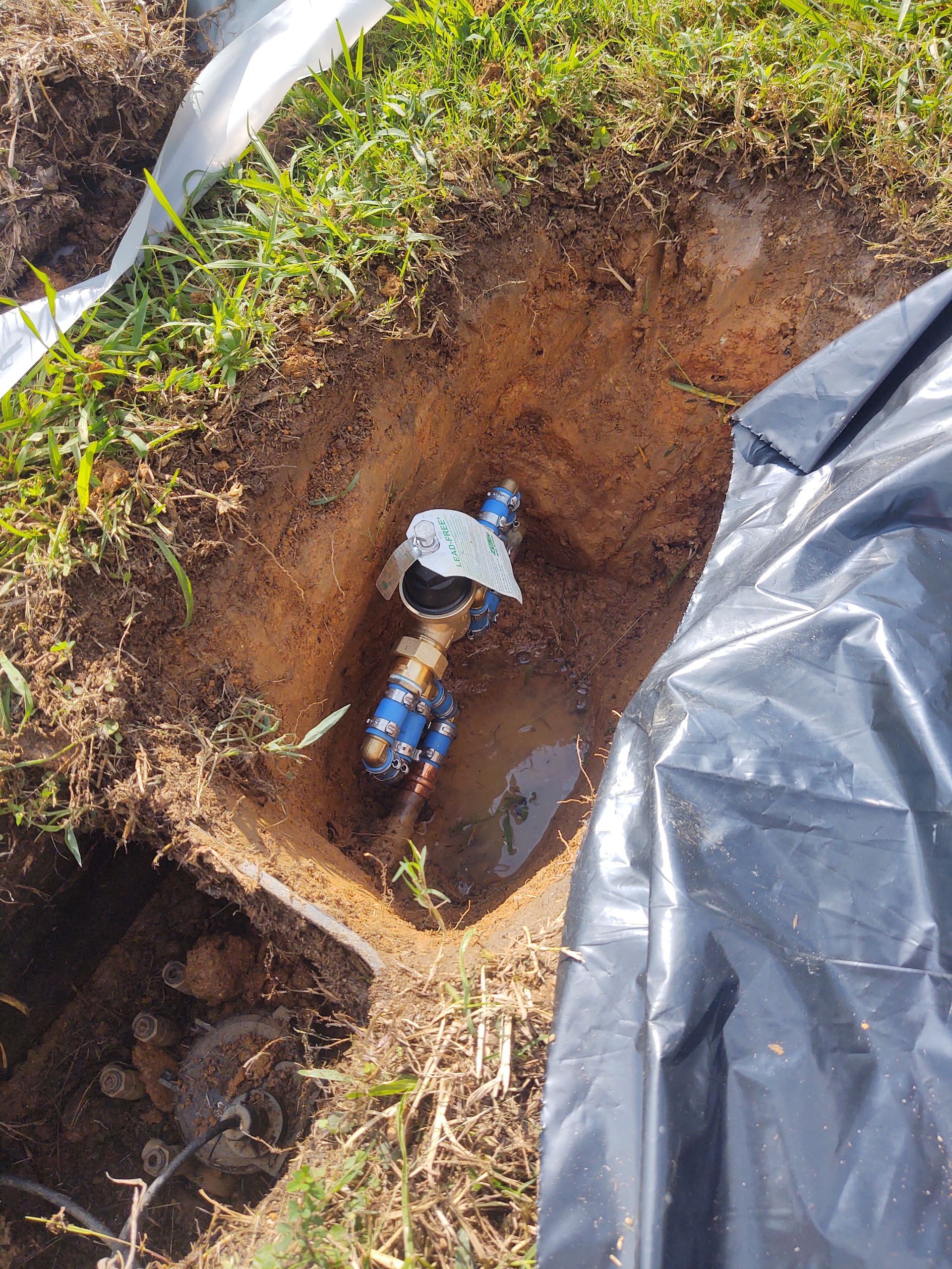 Hole in the ground with water meter, brass pipes, and blue fittings. Surrounding grass and black plastic.