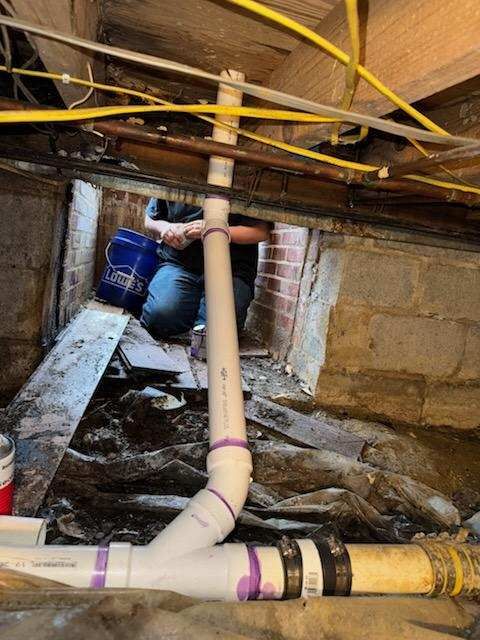 Plumber in crawl space repairs pipe; white and purple PVC pipes with a black fitting, yellow electrical wires overhead.