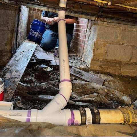 Person installing white PVC pipes in a crawl space with brick and concrete.