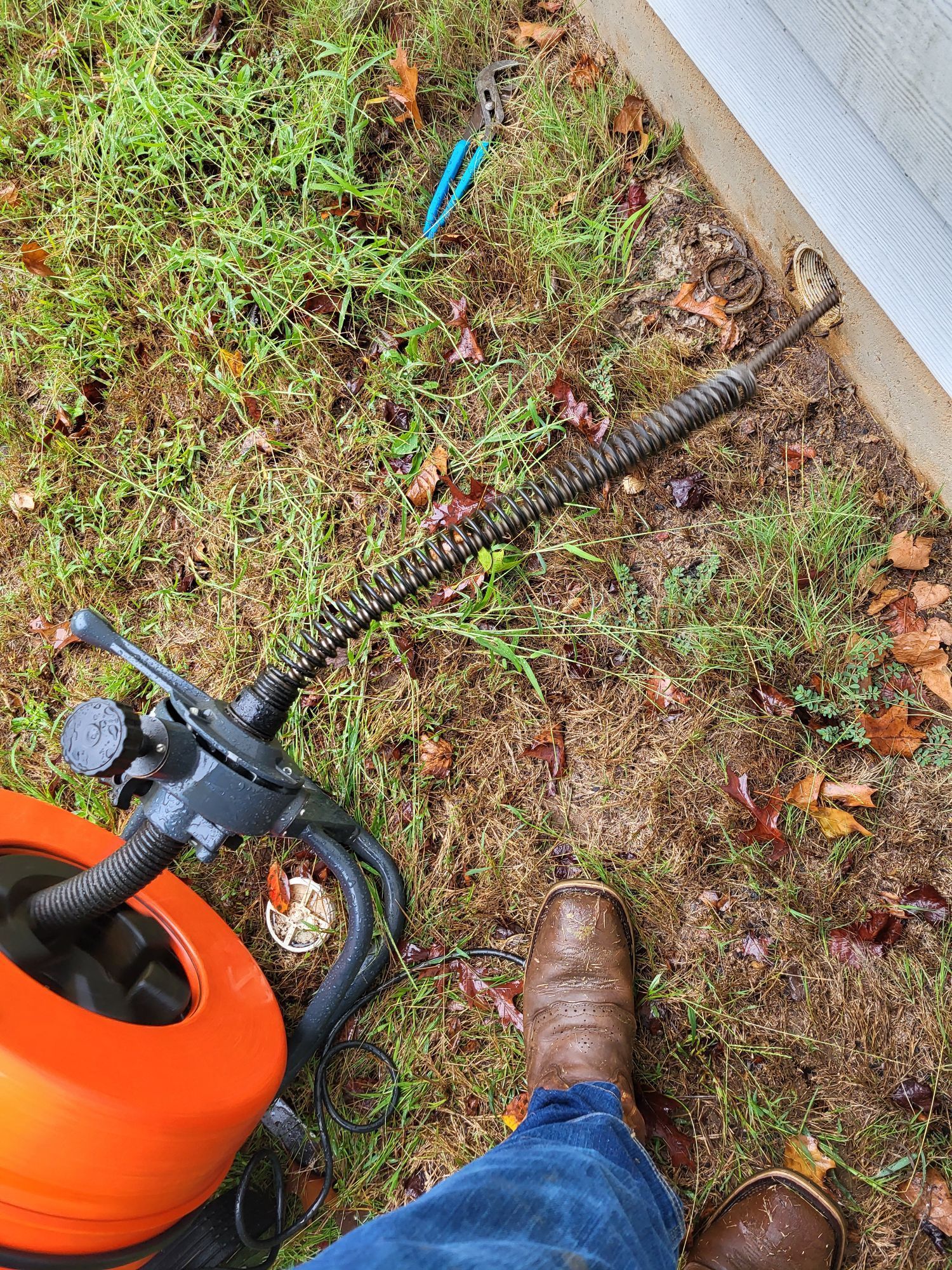 Plumber's snake tool with a coiled cable, next to a boot and orange equipment on wet grass.
