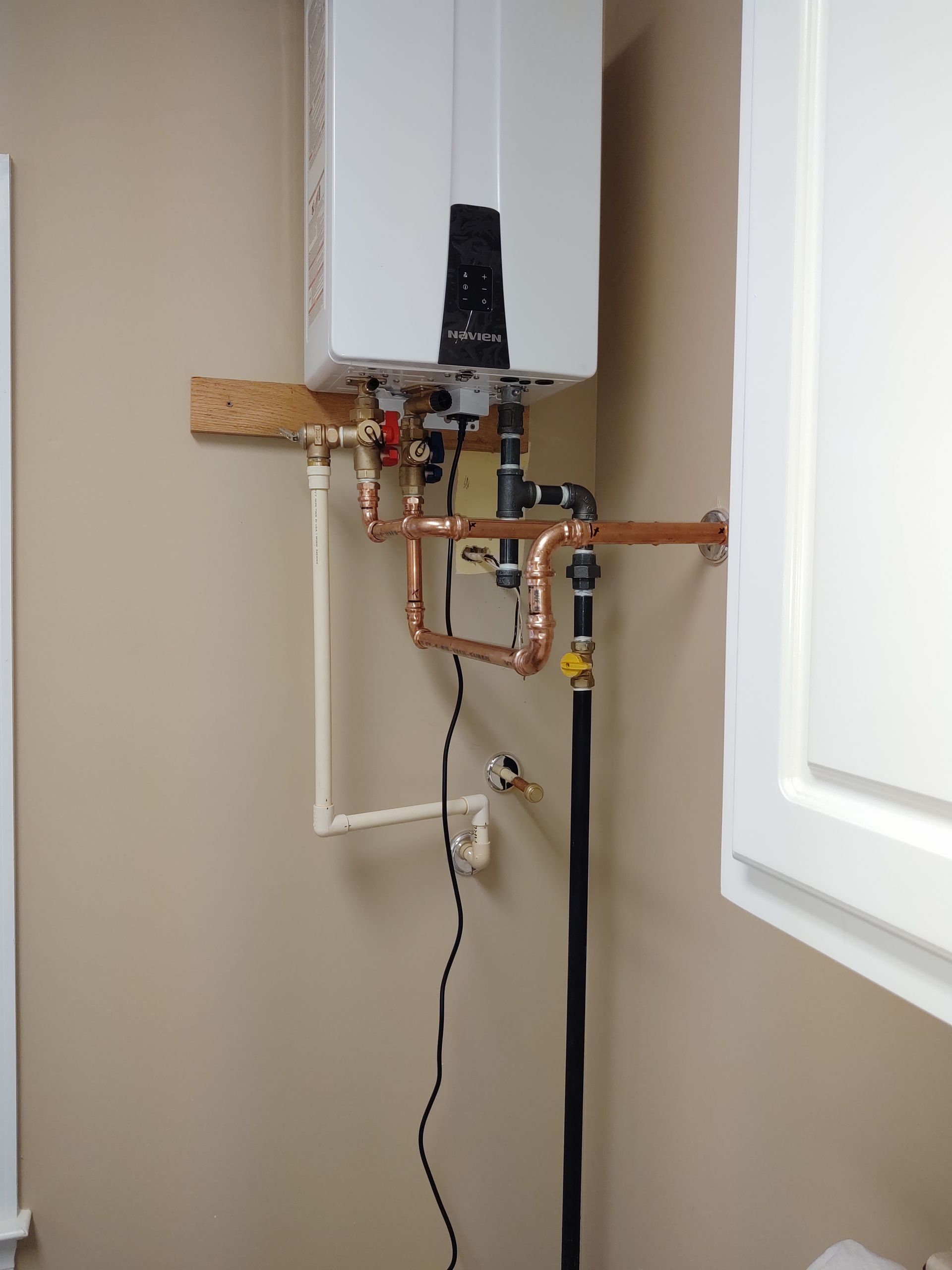 White water heater on a beige wall with copper pipes, wood support, and black gas line near white cabinet.