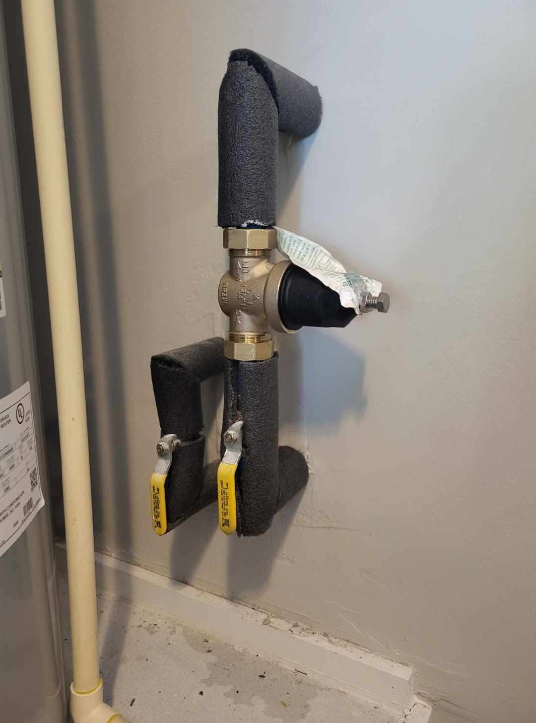 Pipes with black insulation and brass fittings on a gray wall, two yellow-handled valves.