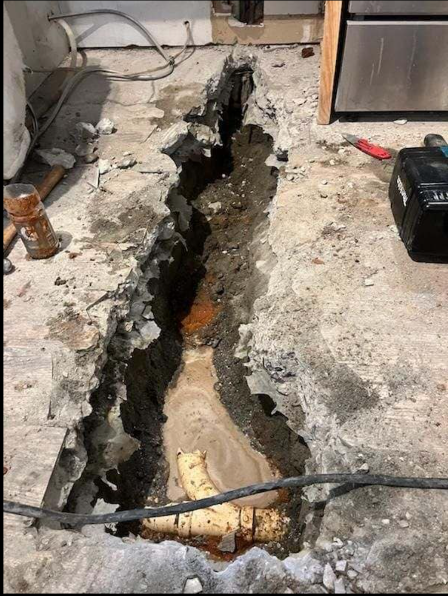 Trench cut in concrete floor, revealing pipes and debris. Construction site.