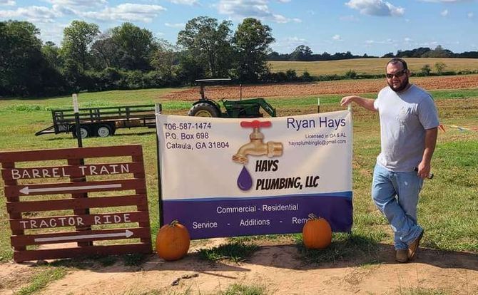 Man holding a banner for Ryan Hays Plumbing, LLC, in a field, with a tractor and sign.
