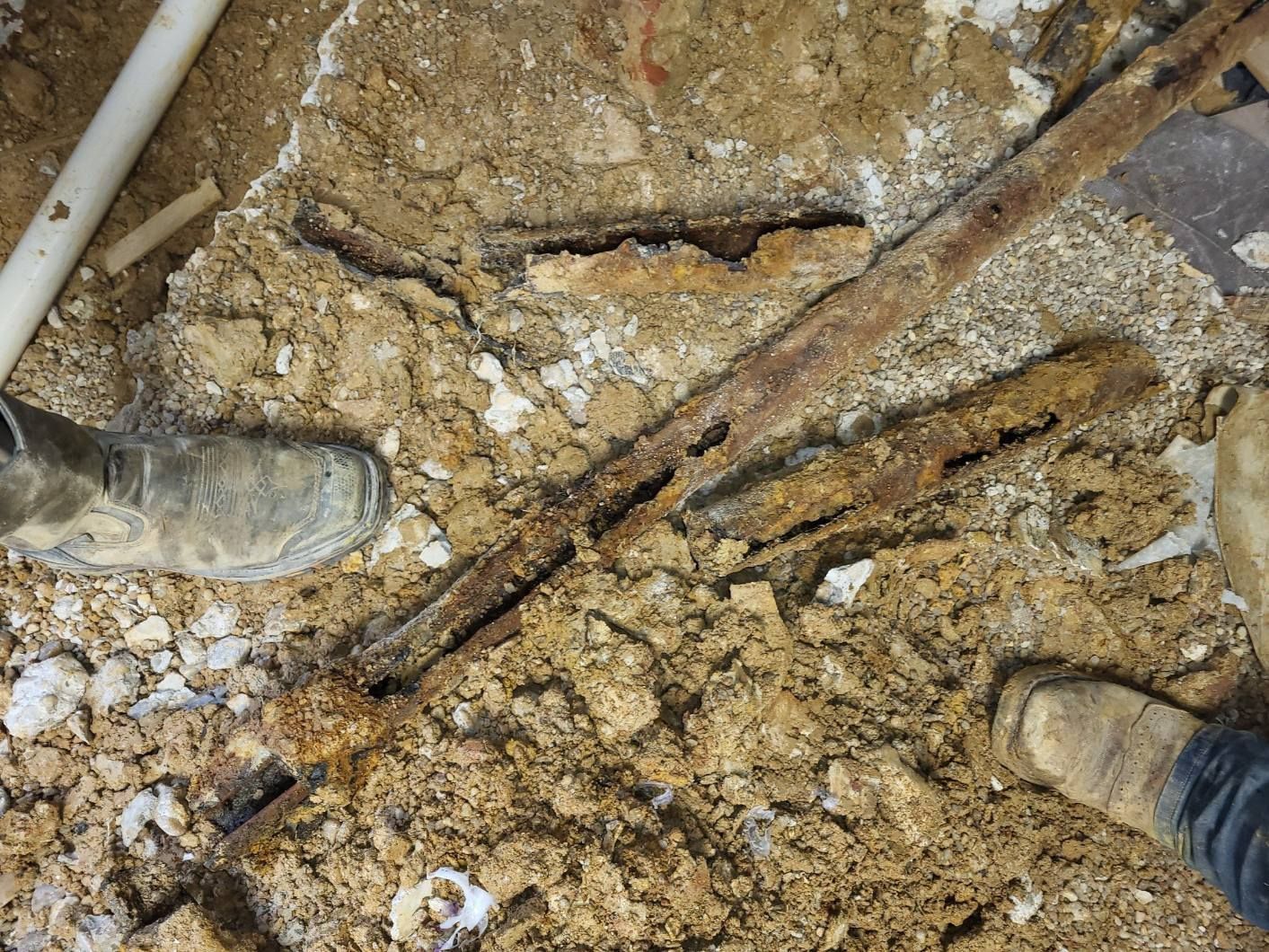 Rusty pipes exposed in dirt, near a white pipe and boots.
