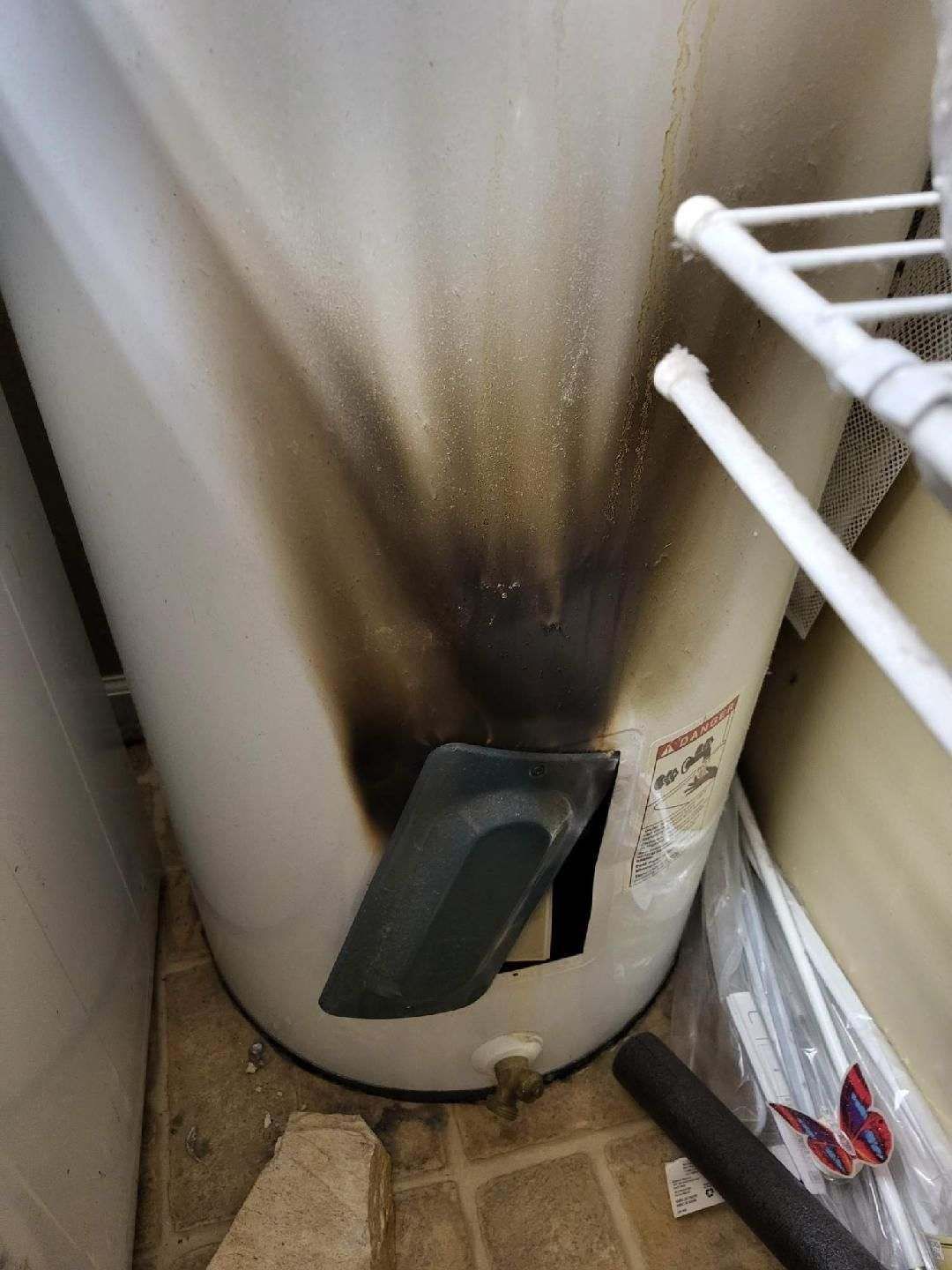 A water heater with significant black burn damage on the side, near the vent, in a utility room.
