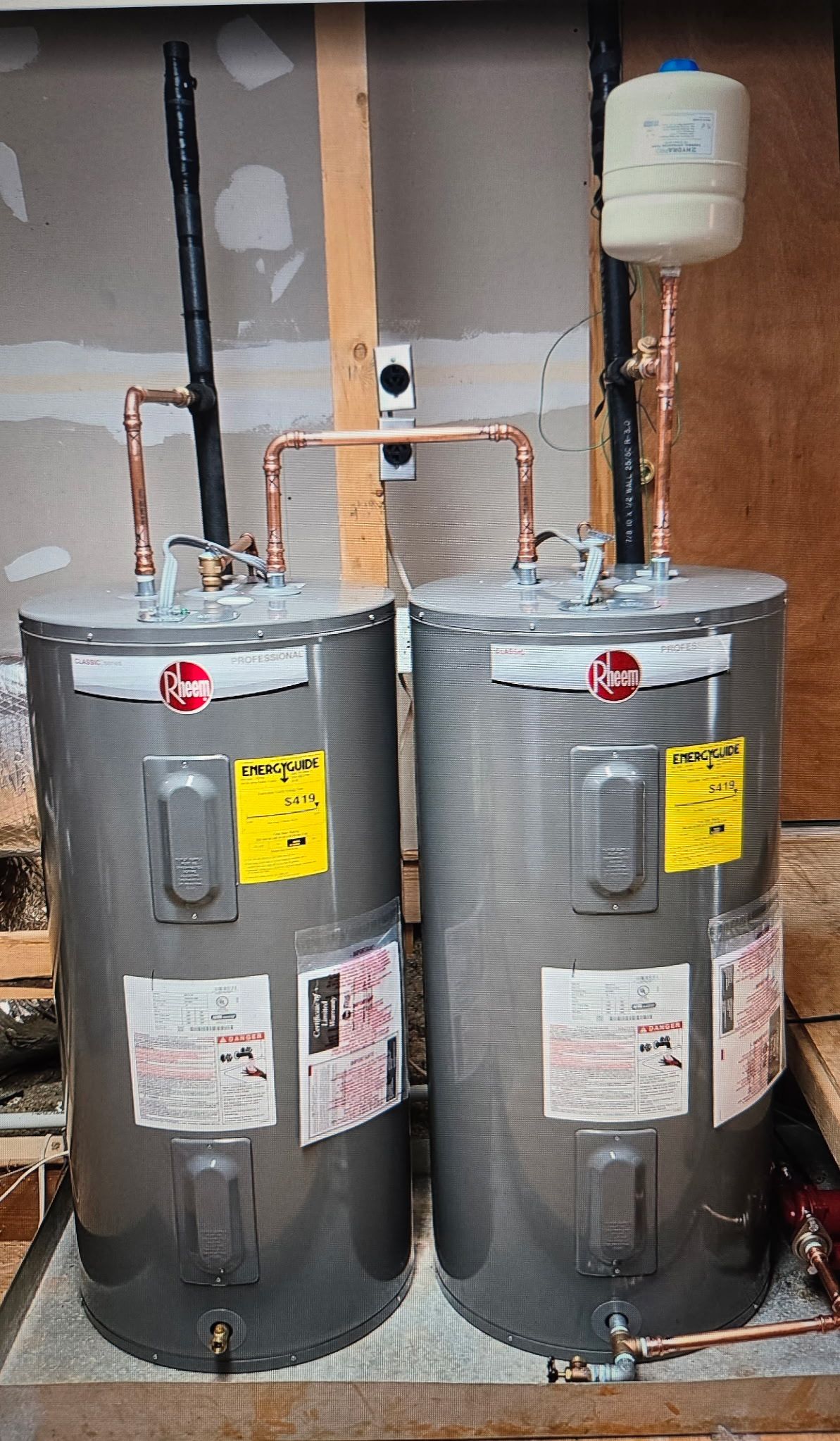 Two Rheem water heaters with copper piping, next to a wooden wall.