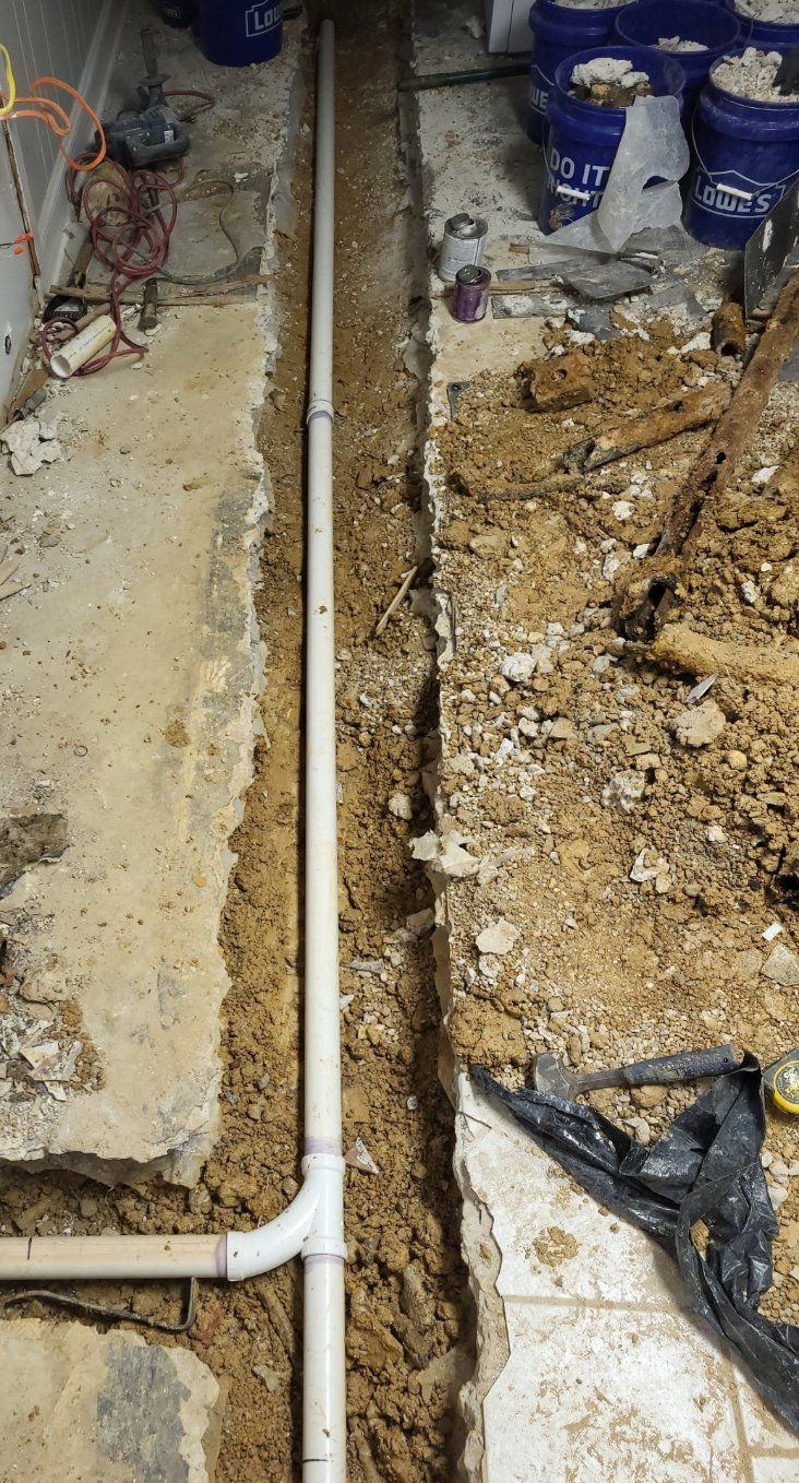 Trench with white PVC pipe, surrounded by dirt, near a concrete floor with buckets in the background.