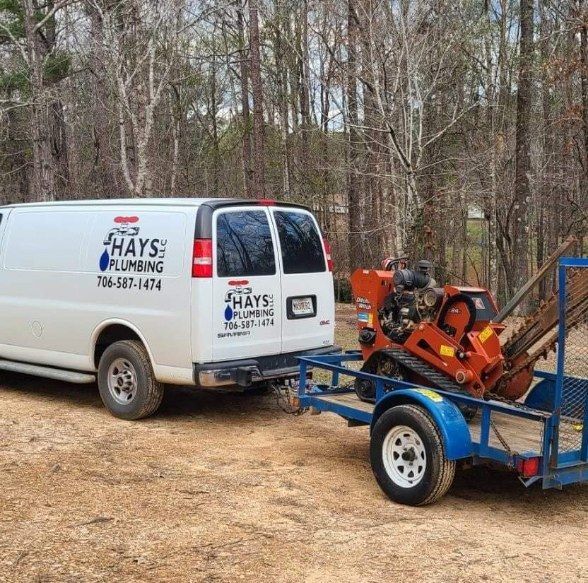 White plumbing van with trailer carrying a ditch witch in wooded area. Hays Plumbing logo visible.