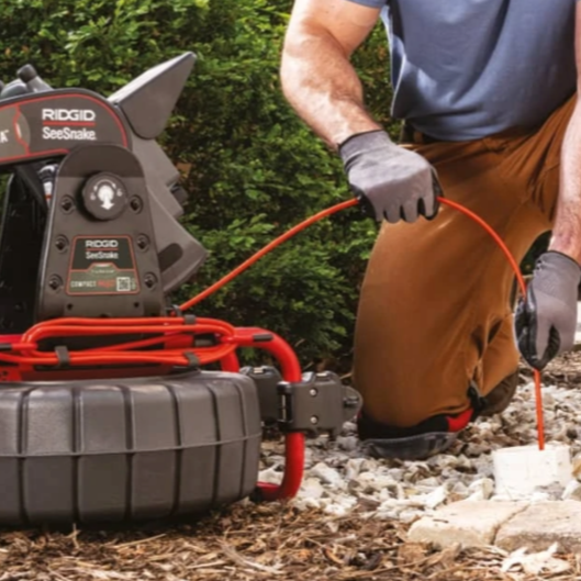 Person using a Ridgid SeeSnake camera to inspect an outdoor drain pipe; red, black, and gray colors; gray gloves.