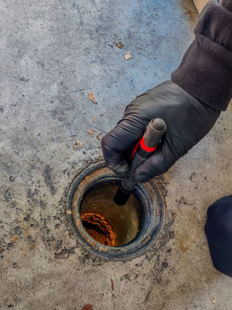 Gloved hand probes rusty drain in concrete floor with red and black tool.