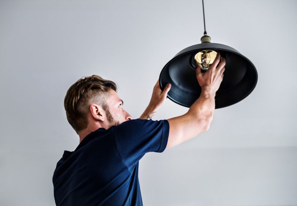 Man Replacing a Light Bulb in a Black Pendant Lamp Hanging From a White Ceiling — Abode Electrical in Tighes Hill, NSW