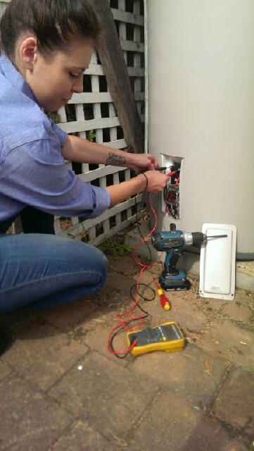 A Woman Testing Wires on a Water Heater Outside, Using a Multimeter and Screwdriver — Abode Electrical in Mayfield, NSW