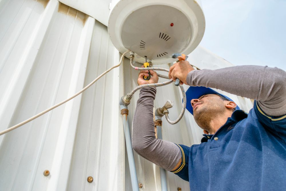 Electrician Repairing a White Water Heater Attached to a Corrugated Metal Wall Outdoor — Abode Electrical in Mayfield, NSW
