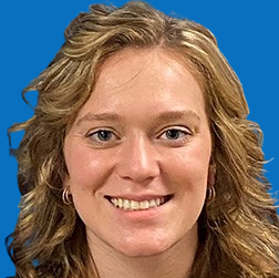 Woman with wavy brown hair smiles at the camera, wearing a blue shirt, against a blue backdrop.