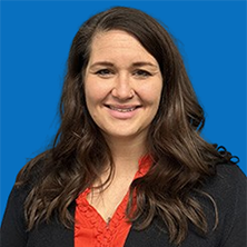 Woman with long brown hair smiles, wearing red shirt and black blazer in front of blue wall.