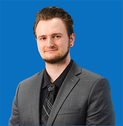 Young man with reddish-brown hair and a goatee, wearing a suit and tie against a blue background.