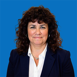 Woman with curly brown hair and pearl necklace smiles. Blue background.