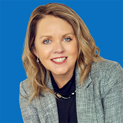 Blond woman with blue eyes smiles, wearing gray blazer, black turtleneck, blue background.