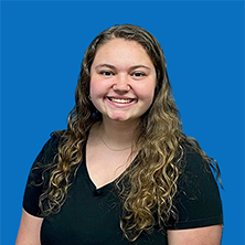Woman with glasses smiles; burgundy shirt, against a blue wall.