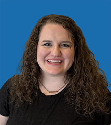Woman with blonde curly hair smiles, wearing a pink blazer and teal top against a blue background.