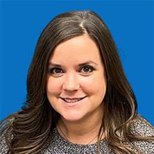 Woman with long brown hair smiles at the camera against a blue background, wearing a gray patterned sweater.