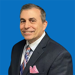Middle-aged man in suit smiles at the camera, blue background.