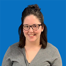 Woman with glasses, smiling, wearing a gray sweater, against a blue wall.
