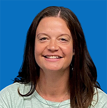 Woman with dark hair smiles at the camera, wearing a light green shirt and a purple pendant, against a blue backdrop.