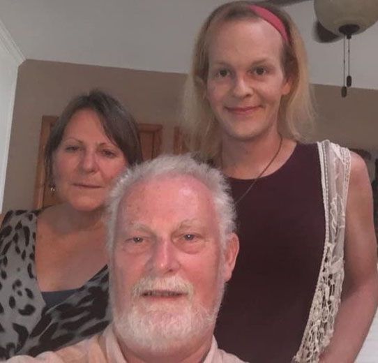 Family smiling for a photo; a man with a white beard, a woman, and a person in a dress and a headband.