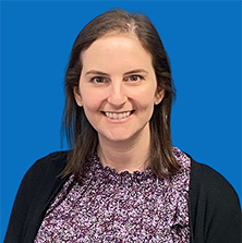Woman with brown hair smiling, wearing a purple floral top and black cardigan in front of a blue wall.