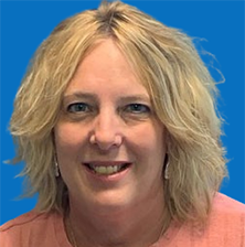 Blonde woman with blue eyes smiles, wearing a pink shirt, in front of a teal background.