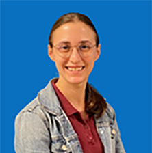 Woman with glasses smiles, wearing a jean jacket and burgundy polo shirt, standing against a teal wall.