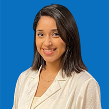 Woman with dark hair smiles, wearing a white blazer, against a blue background.