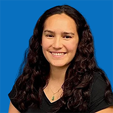 Woman with long dark curly hair smiles at the camera, wearing a black shirt and a necklace; blue background.