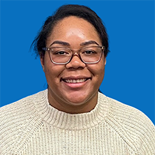 Woman with glasses smiles at the camera against a blue backdrop, wearing a cream-colored sweater.