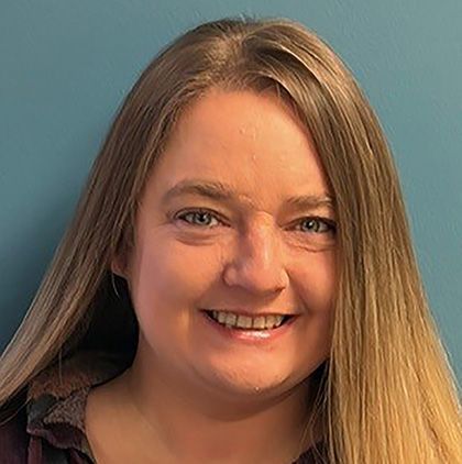 Woman with blonde hair and blue eyes, looking towards the camera, against a blue background.