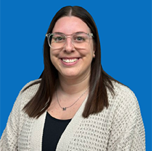 Woman with glasses smiles, wearing a cardigan and necklace, against a blue background.