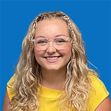 Woman with blonde wavy hair, glasses, and a yellow top smiles at the camera against a blue background.