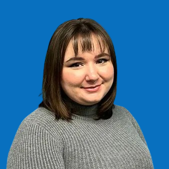 Woman with brown hair and bangs wearing a grey turtleneck smiles at the camera, with a blue wall background.