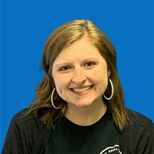 Smiling woman with light skin, wearing earrings and a black jacket, against a blue background.