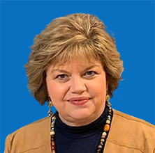 Woman with short blonde hair wearing a beige jacket and beaded necklace, against a blue background.